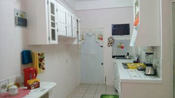 A small kitchen with white cabinets, a refrigerator, a sink, and a stove. There is a window with a curtain and a door with a decorative wreath.