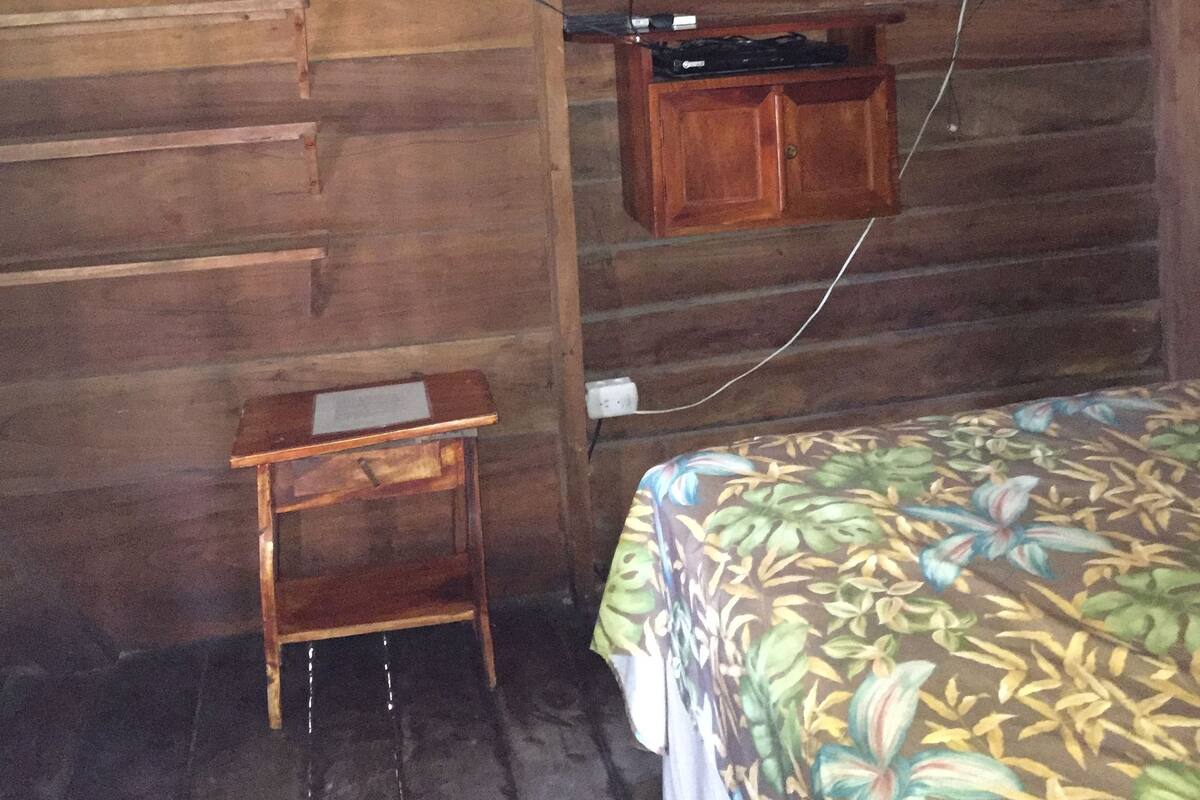 A wooden room with a bed, a small wooden table, a television mounted on the wall, and a wall-mounted fan.
