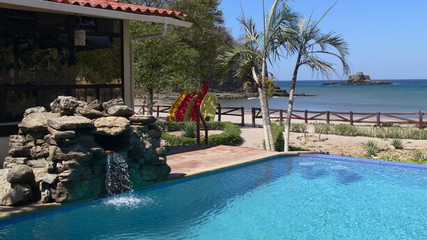 Una piscina con una cascada rocosa, vistas a la playa y un edificio con techo de tejas rojas.