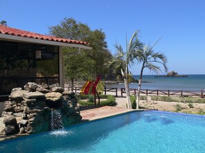 A pool with a rocky waterfall feature, a beach view, and a building with a red-tiled roof. - Marsella Beachfront B&B (San Juan del Sur)
