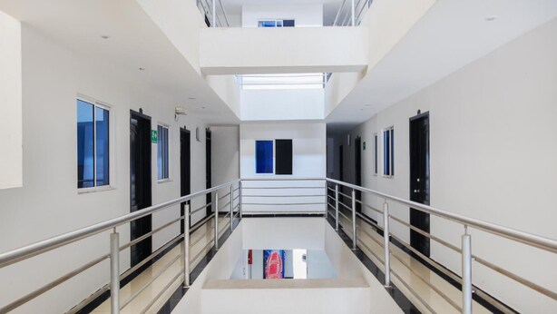 Un pasillo moderno de hotel con una escalera central, barandales de metal y un ascensor de vidrio.