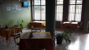 Una habitación amplia con varias mesas y sillas, ventanas grandes con barrotes y un televisor montado en la pared.