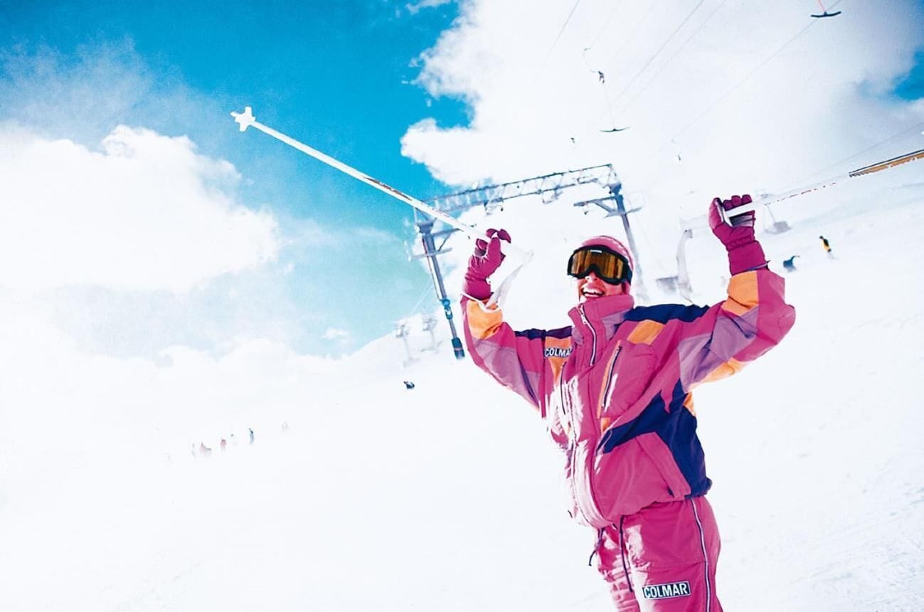 A person in a pink and blue ski suit with a 'COLMAR' label, standing on a snowy slope with ski lifts in the background.