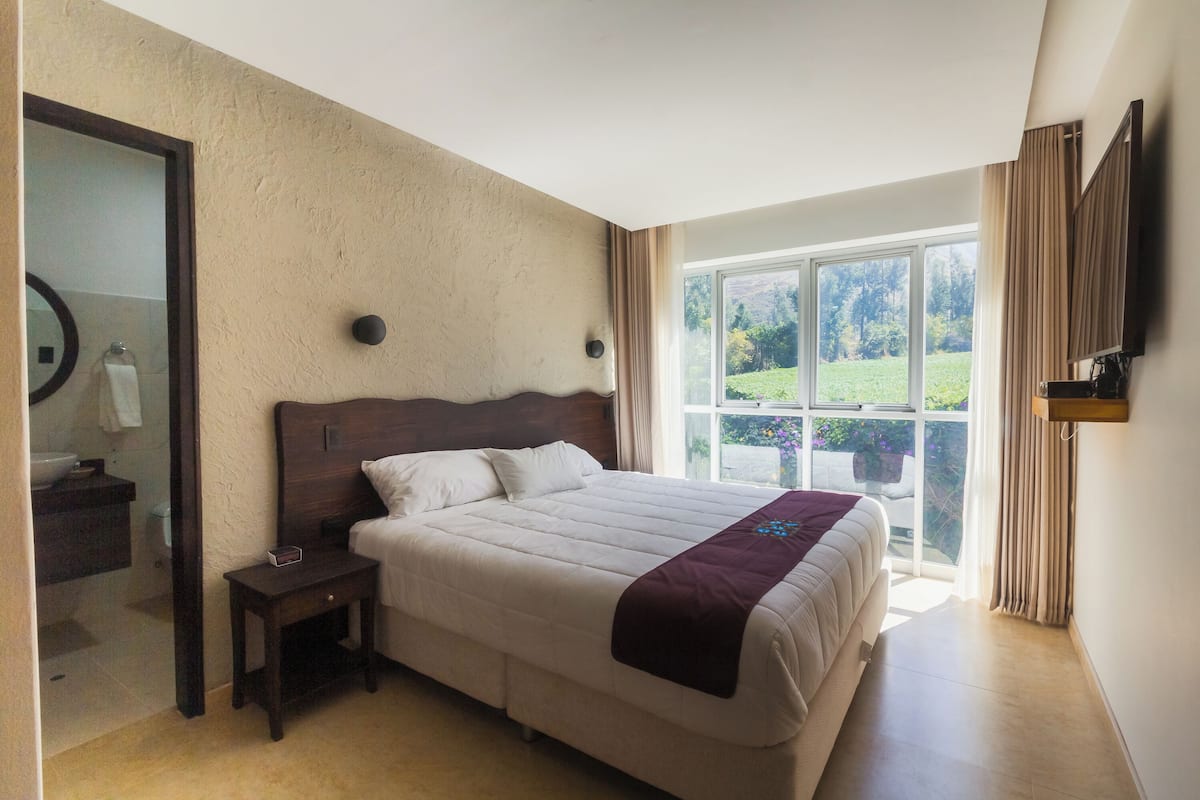 Hotelkamer met een groot bed, nachtkastje, spiegel en raam met uitzicht op groen.