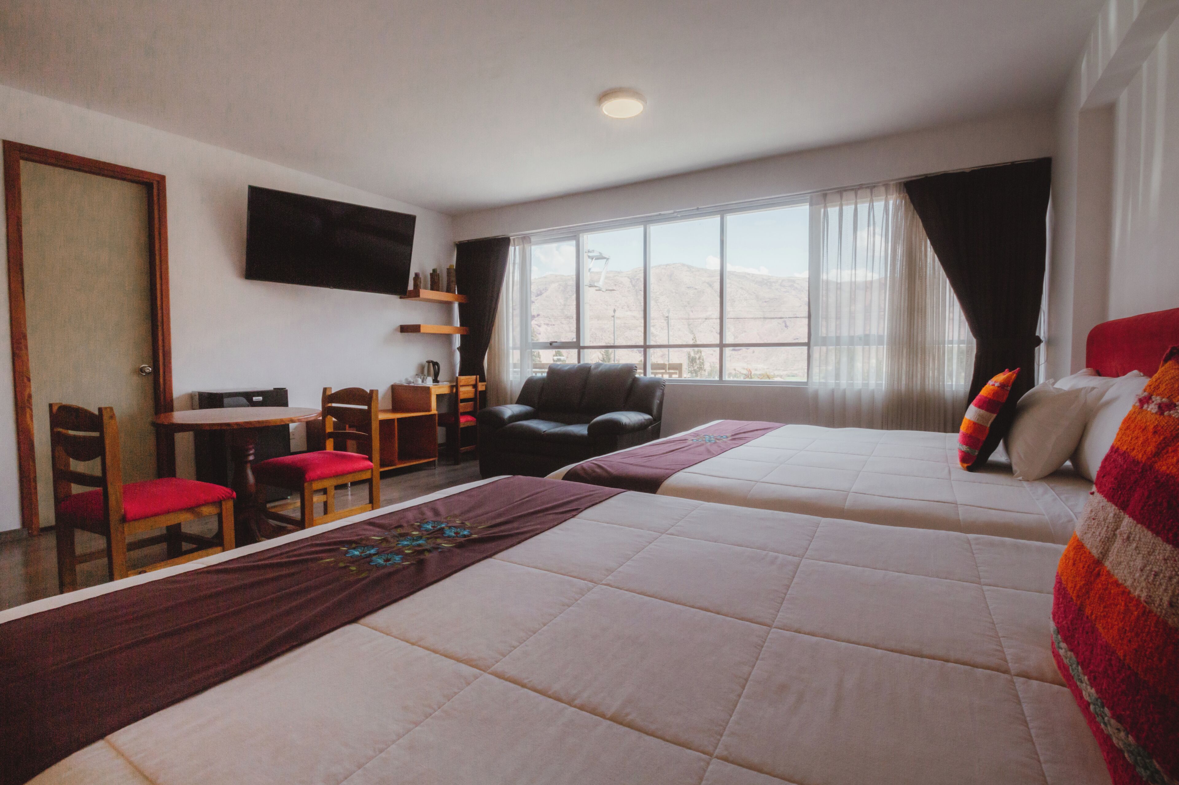 Habitación de hotel con dos camas, un escritorio, sillas, televisor y un amplio ventanal con vistas a la montaña.