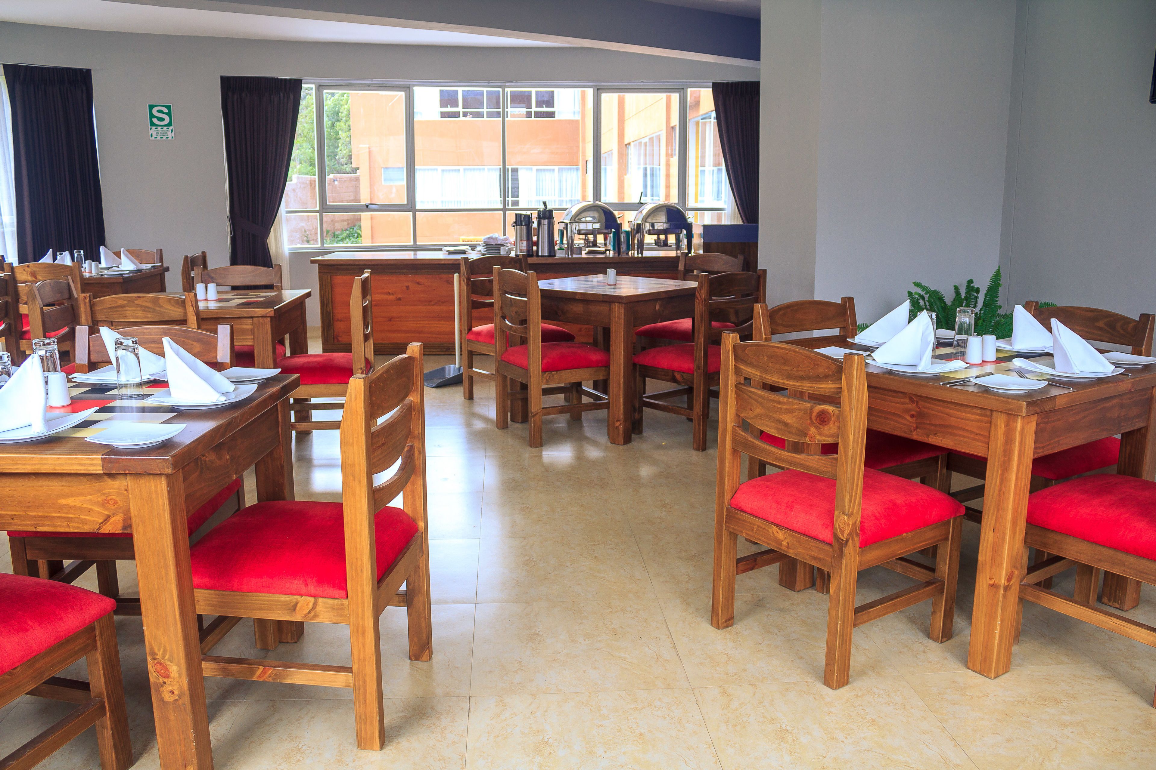 Un comedor con mesas de madera y sillas rojas con cojines, una ventana con cortinas y vistas a edificios.