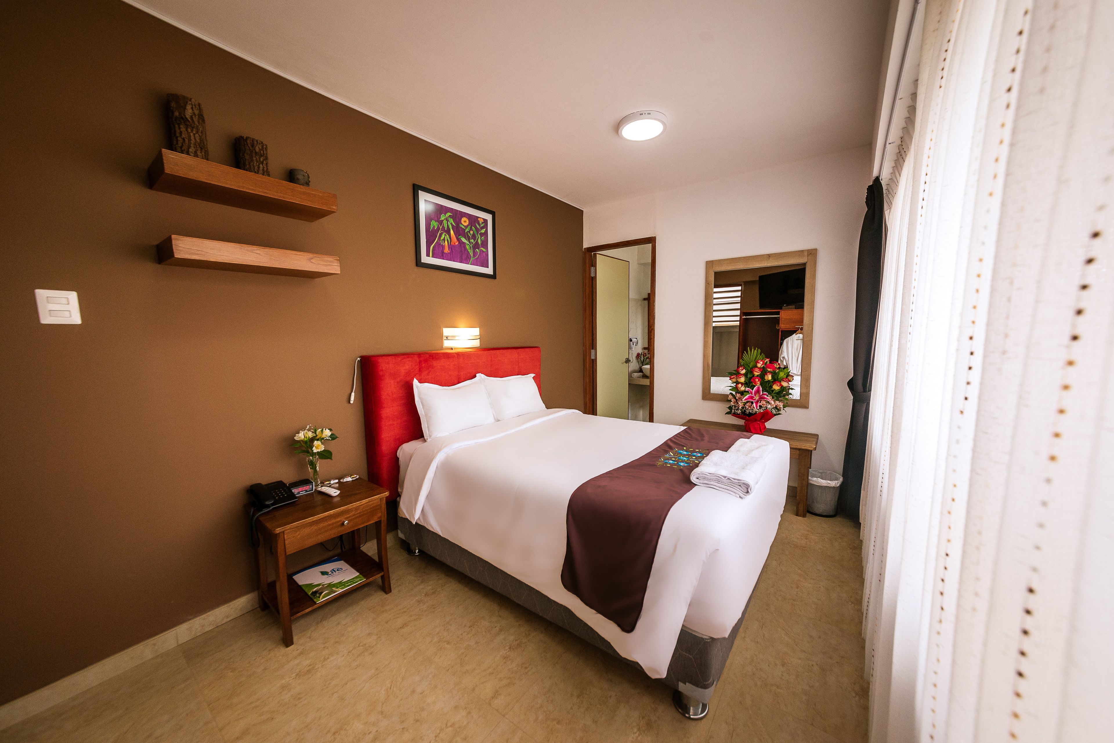 A hotel room with a large bed, a nightstand, a wooden shelf with decorative items, a mirror, and a window with curtains.