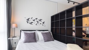 Un lit bien fait, avec une literie blanche et violette, une étagère fixée au mur et un motif décoratif représentant des oiseaux.