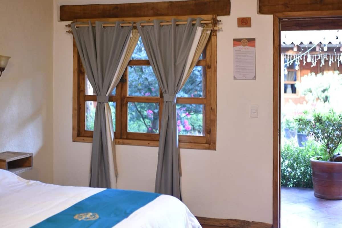 A room with a wooden ceiling, a bed with a teal bedspread, two windows with curtains, and a door leading outside.
