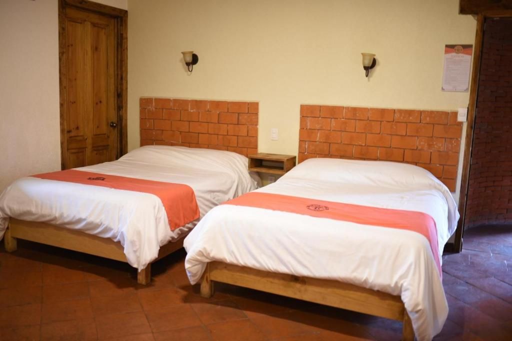 Dos camas individuales con cabeceros de madera y ropa de cama blanca, una puerta de madera y una pared de ladrillo con dos apliques.