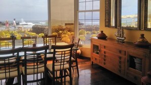 A dining area with large windows overlooking a rocky coastline and cacti. There is a wooden cabinet with decorative items and a mirror on the wall. - Mision Costa (La Oliva)