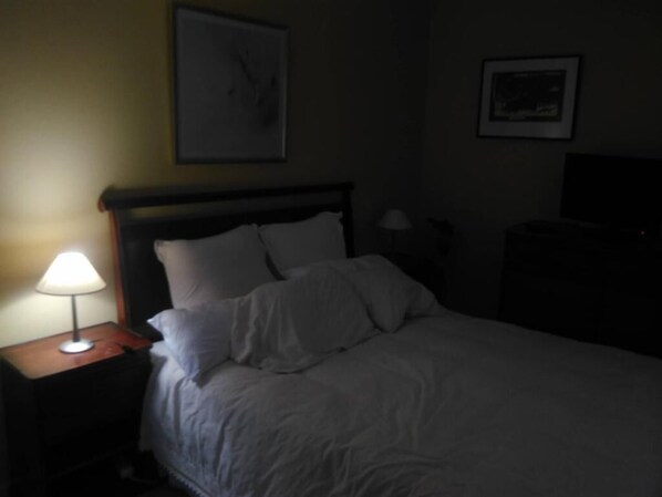 A dimly lit hotel room with a bed, a nightstand with a lamp, and a wall-mounted TV. - Mision Costa (La Oliva)