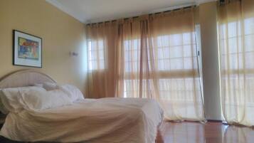Un dormitorio con cama, cortinas color crema y un cuadro en la pared.