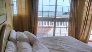 A bed with white linens and a view of the ocean through a window with a white railing. - Mision Costa (La Oliva)