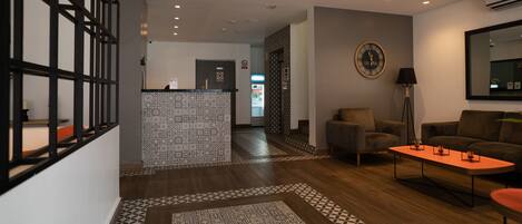 A modern lobby with a reception desk, patterned floor tiles, a sofa, a coffee table, and a wall-mounted clock.