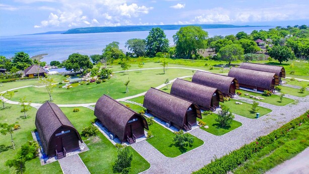 A resort with multiple rustic, barrel-shaped cabins arranged in a semi-circle, surrounded by well-maintained lawns and a view of a large body of water. - Samawa Seaside Resort (Sumbawa Besar)