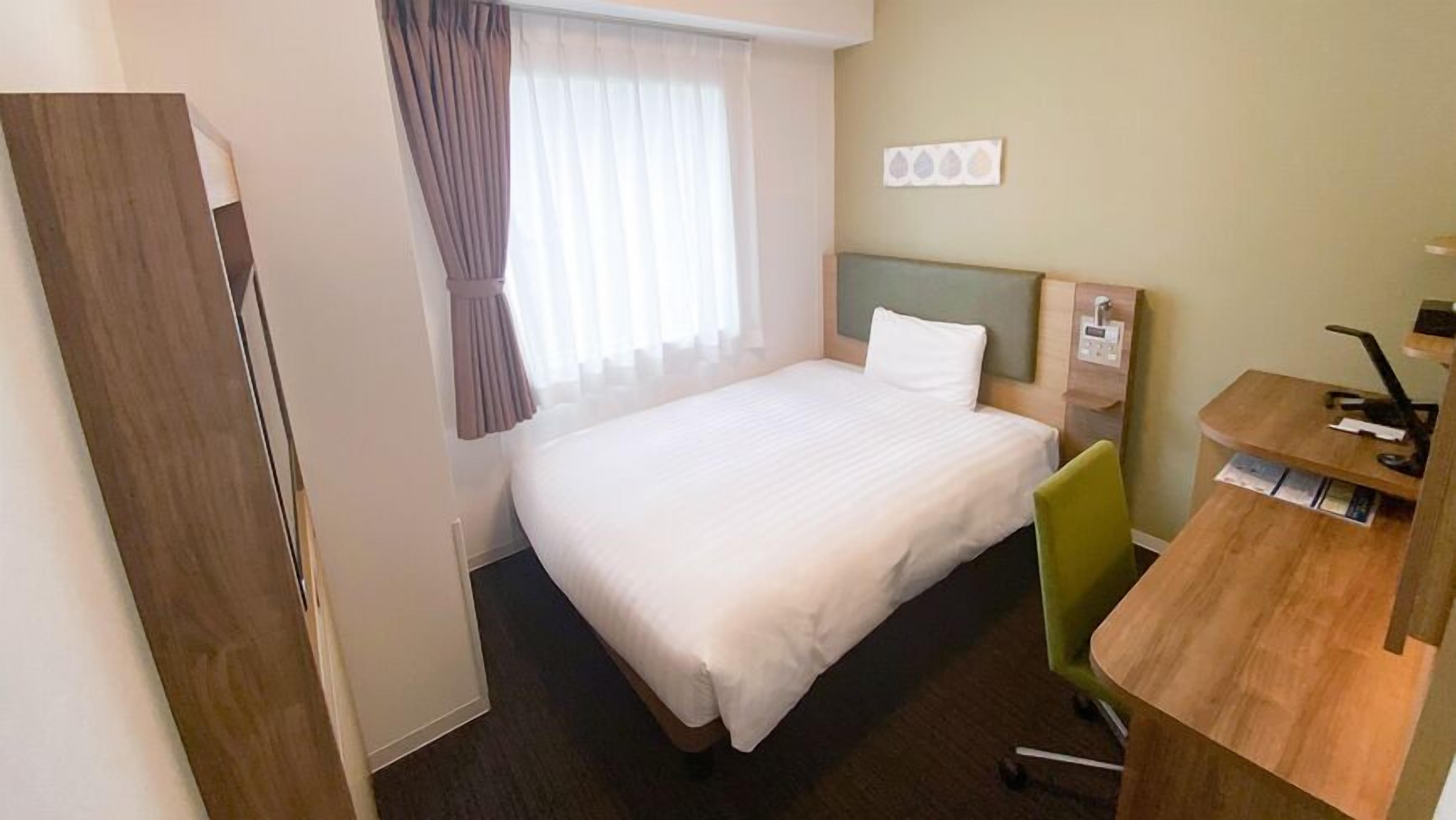 A hotel room with a bed, a desk with a chair, a window with curtains, and a wall-mounted control panel.
