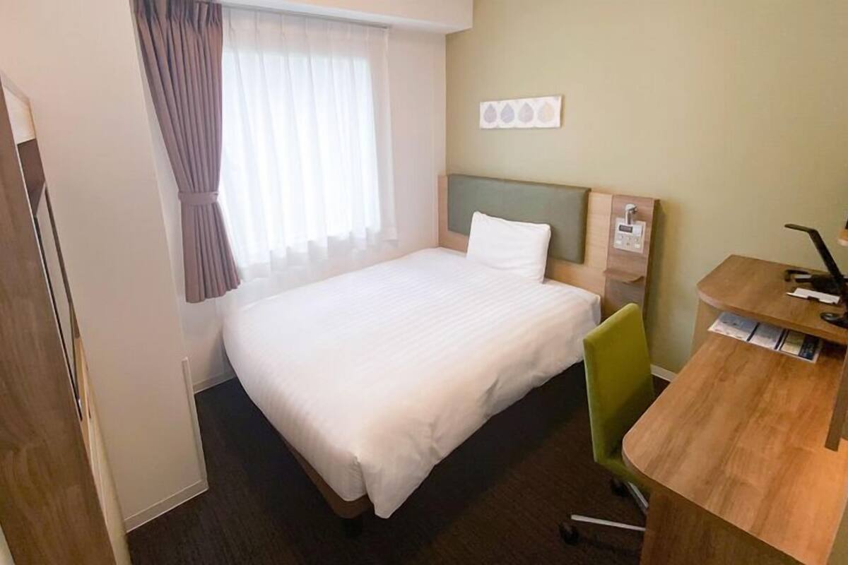 Habitació d'hotel amb un llit, una taula de treball amb una cadira, una finestra amb cortines i un panell de control fixat a la paret.
