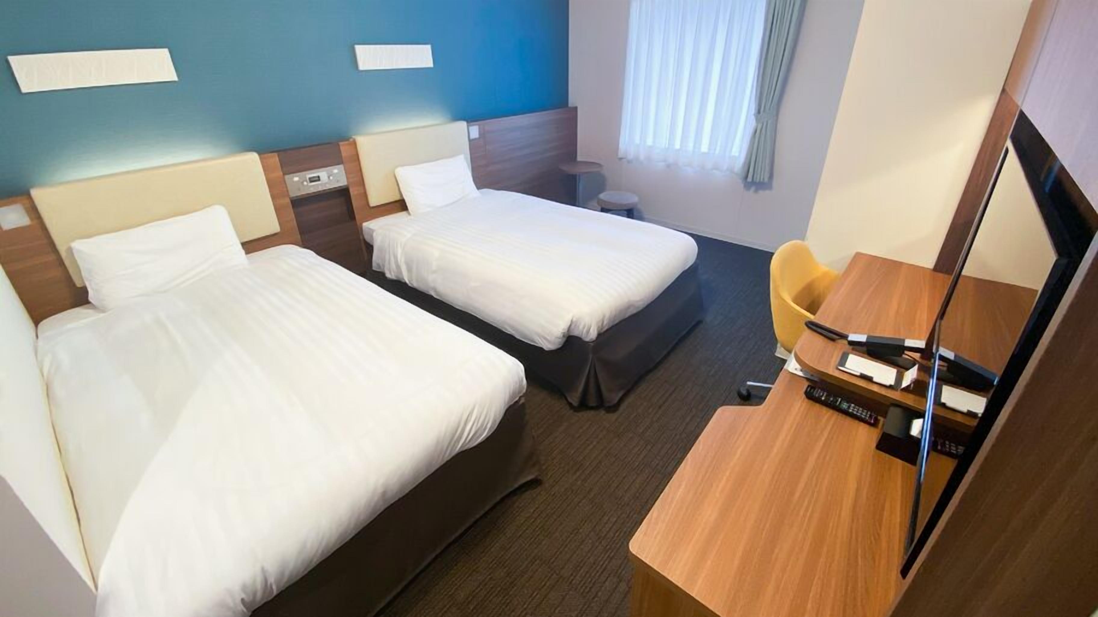A hotel room with two beds, a desk with a chair, a small table, a mirror, and a window with curtains.
