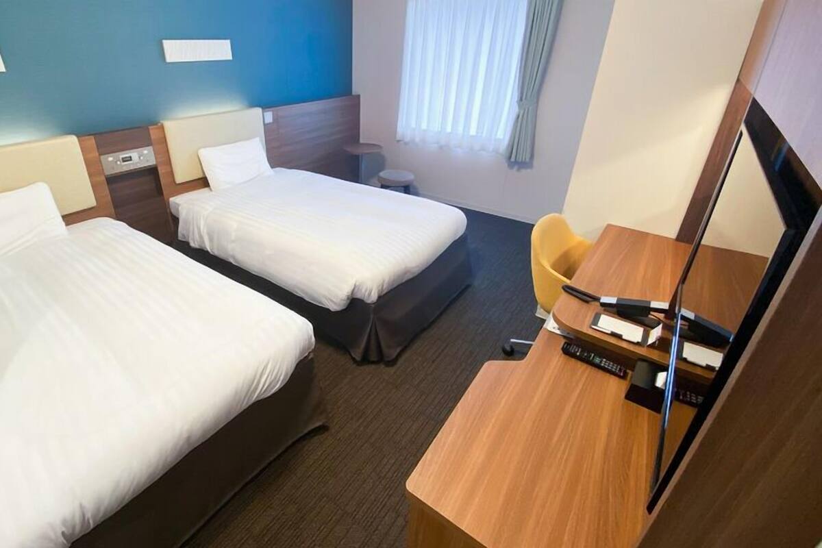 Habitació d'hotel amb dos llits, un escriptori amb cadira, una taula petita, un mirall i una finestra amb cortines.
