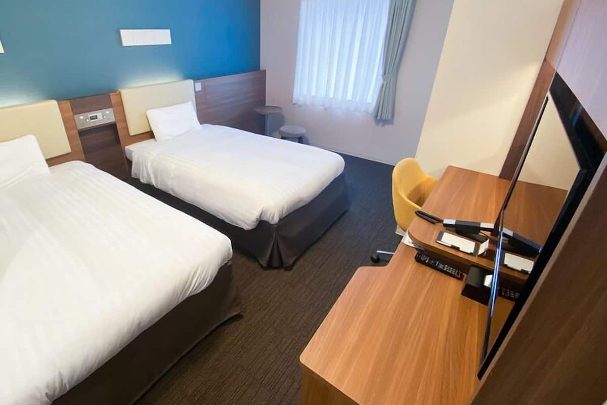 A hotel room with two beds, a desk with a chair, a small table, a mirror, and a window with curtains.