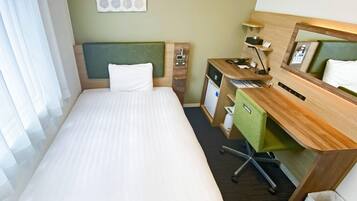Una habitación de hotel con cama, escritorio con silla, espejo, lámpara y un pequeño cubo de basura.