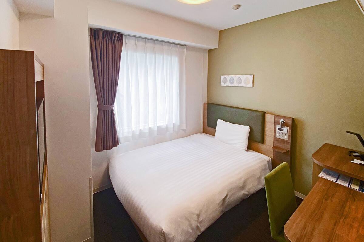 Habitació d'hotel amb un llit, una taula de treball, una cadira i una finestra amb cortines.