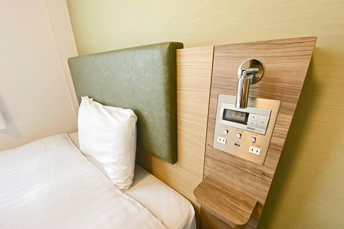 Et hotelværelse med en seng, en sengegavl og et kontrolpanel med diverse knapper og et digitalt display.