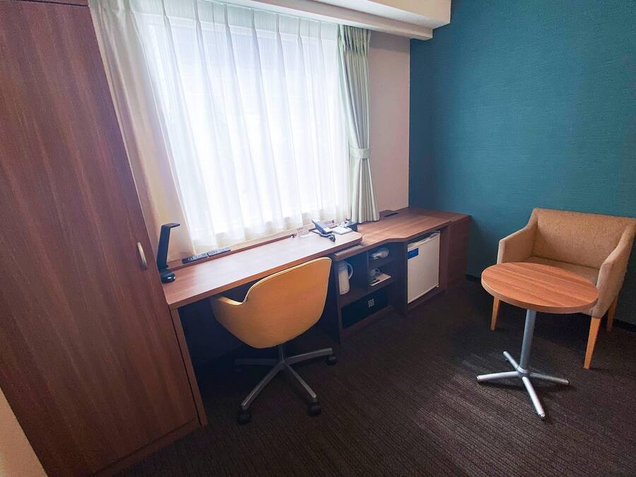 A hotel room with a wooden desk, a chair, a small table, and a wardrobe.
