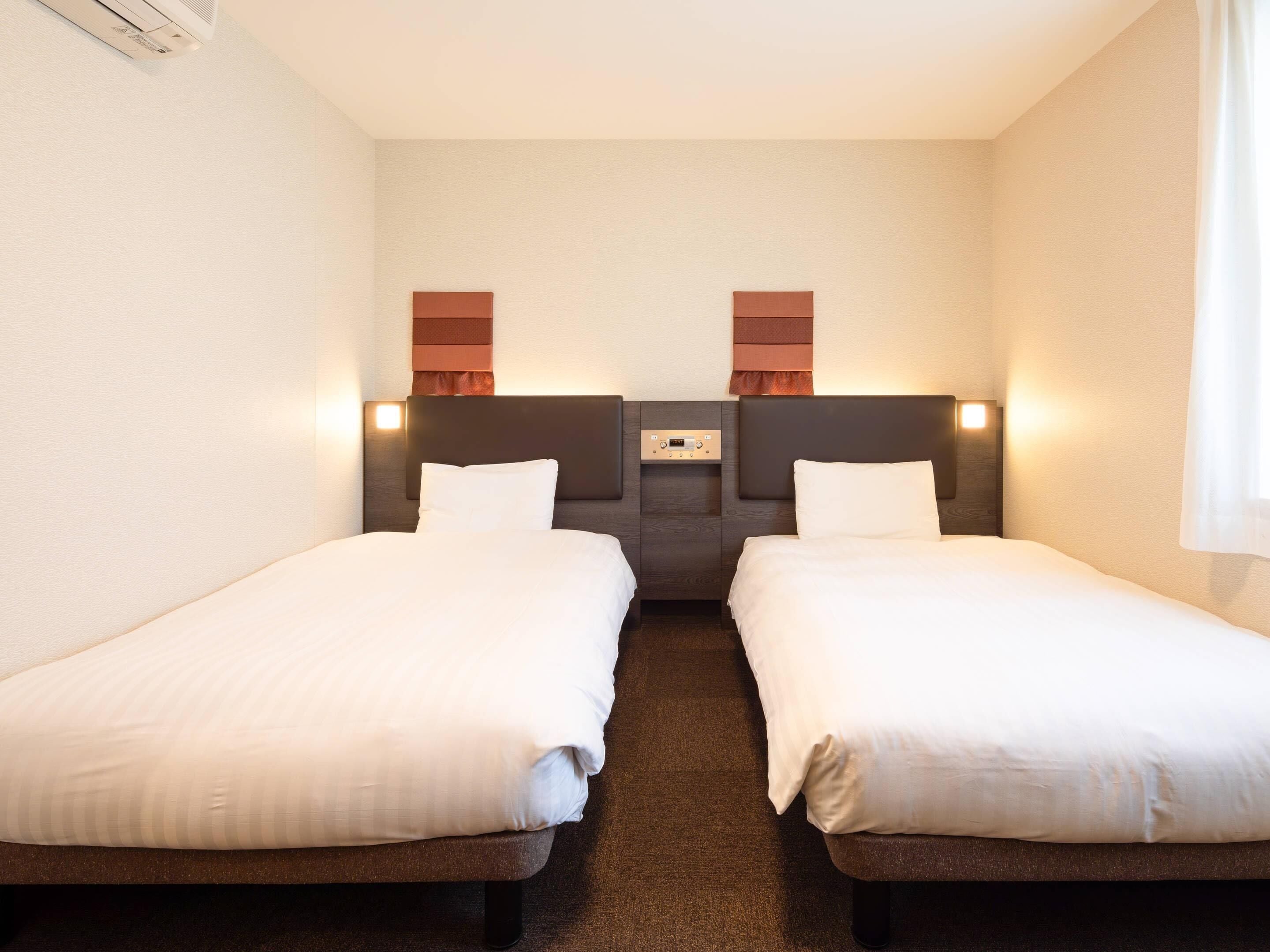 A hotel room with two single beds, a small seating area, and a wall-mounted control panel.