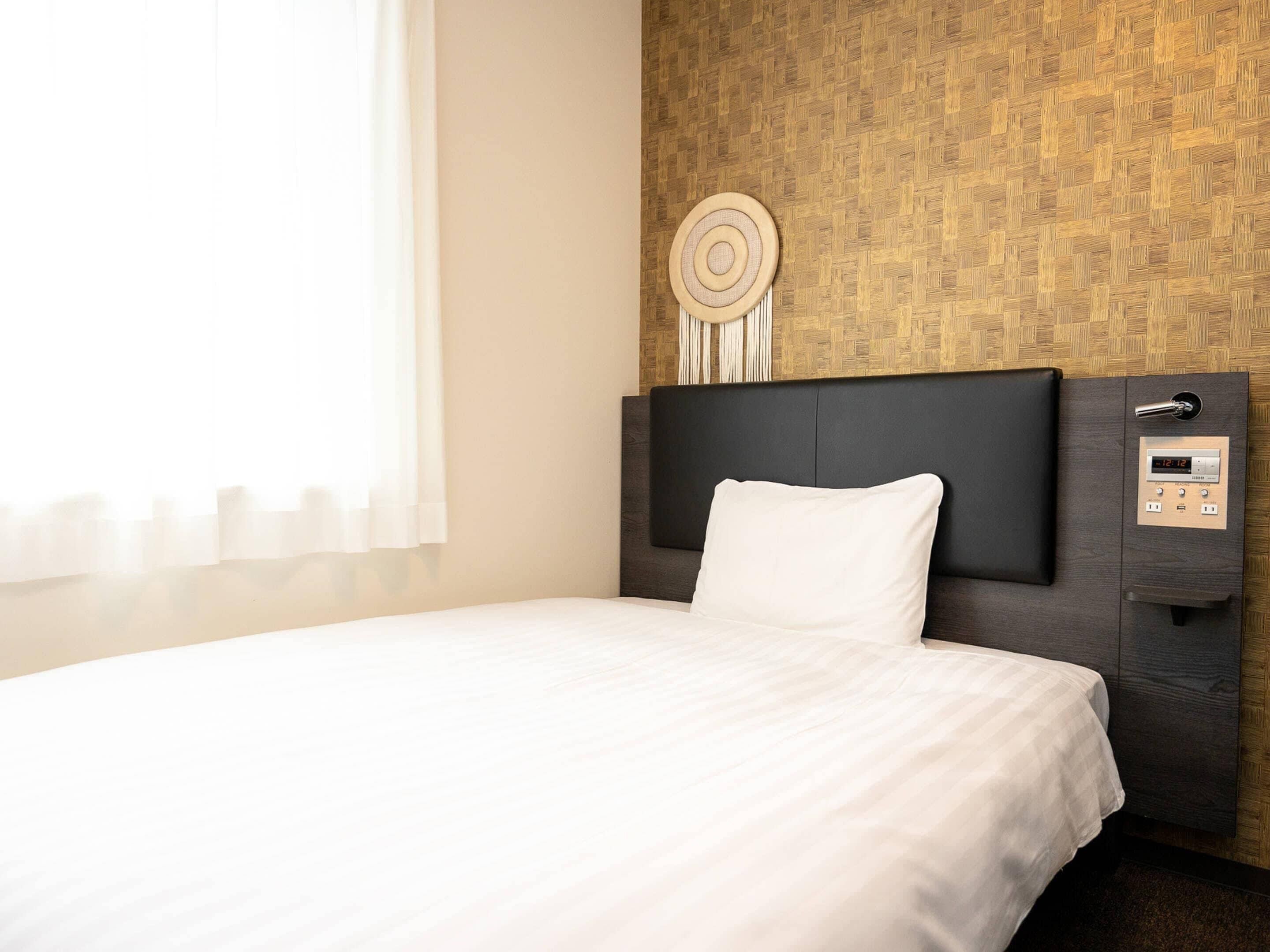 A hotel room with a bed, a headboard with a control panel, a wall-mounted decorative item, and a window with curtains.
