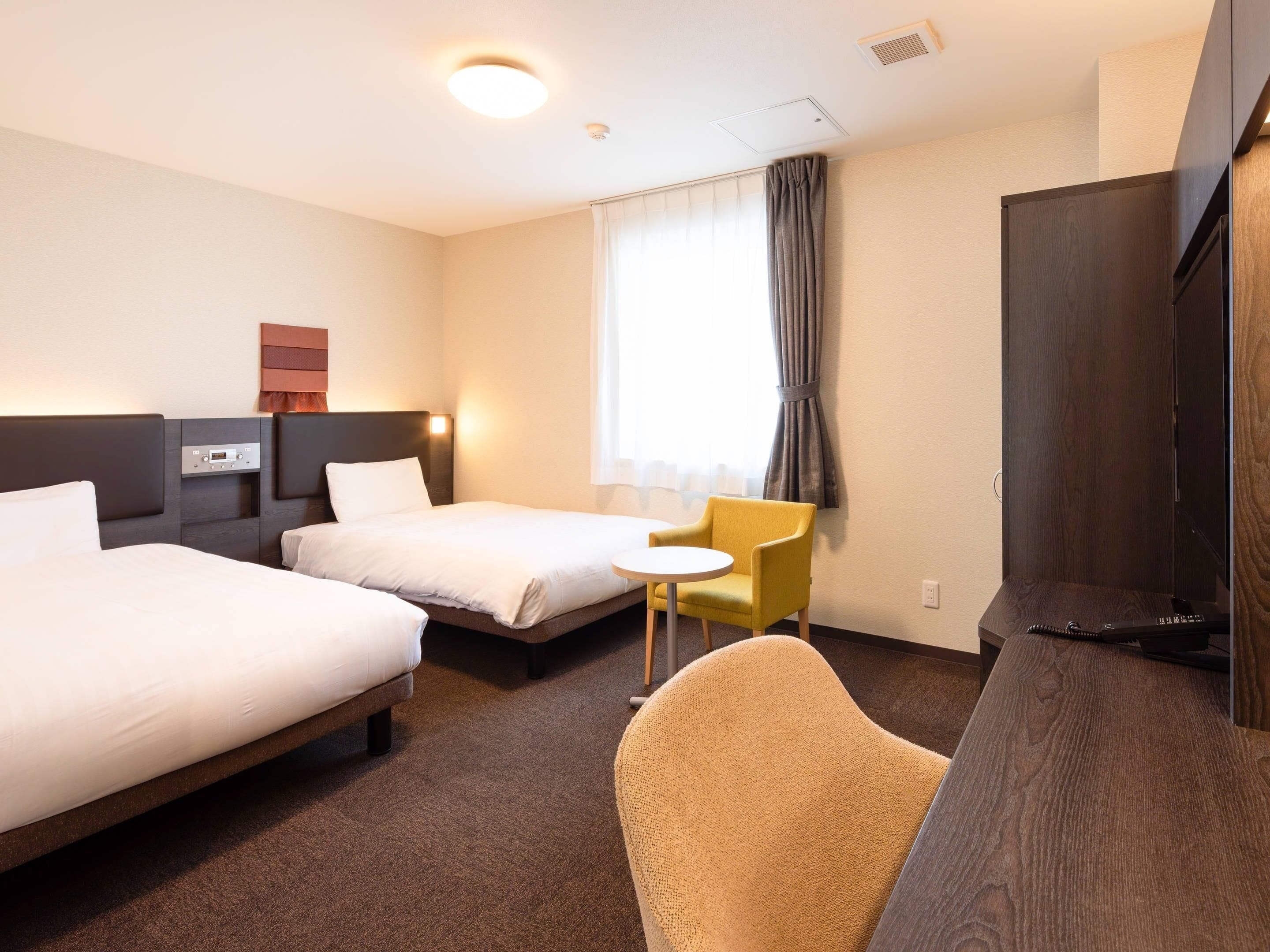 A hotel room with two beds, a small table, a chair, a TV, and a window with curtains.