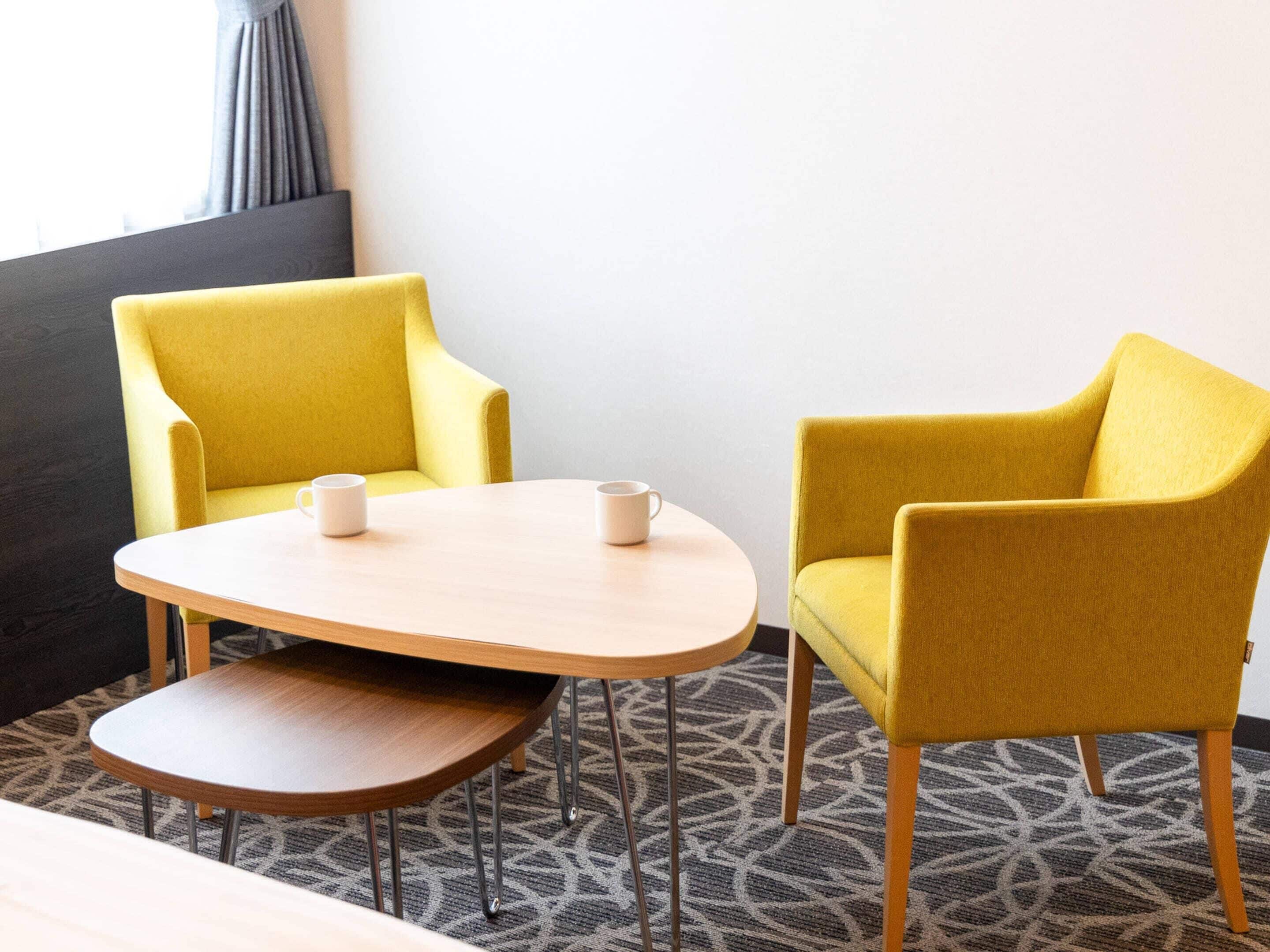 A hotel room with a yellow armchair, a wooden side table, and a small round table with two white mugs.