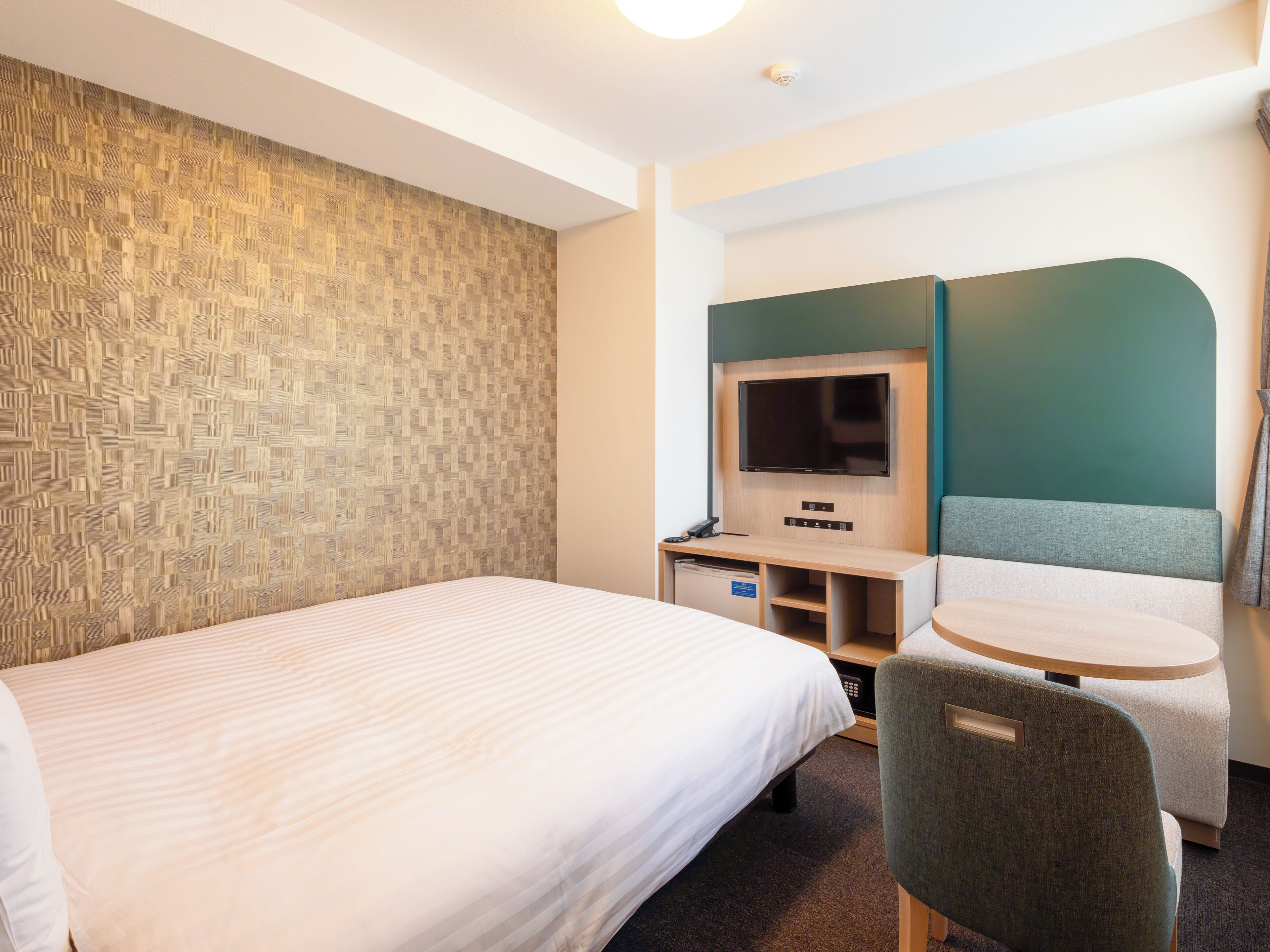 Quarto de hotel com cama, escrivaninha com televisão, uma cadeira e uma pequena mesa.