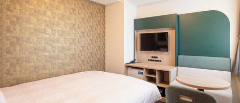 Quarto de hotel com cama, escrivaninha com televisão, uma cadeira e uma pequena mesa.