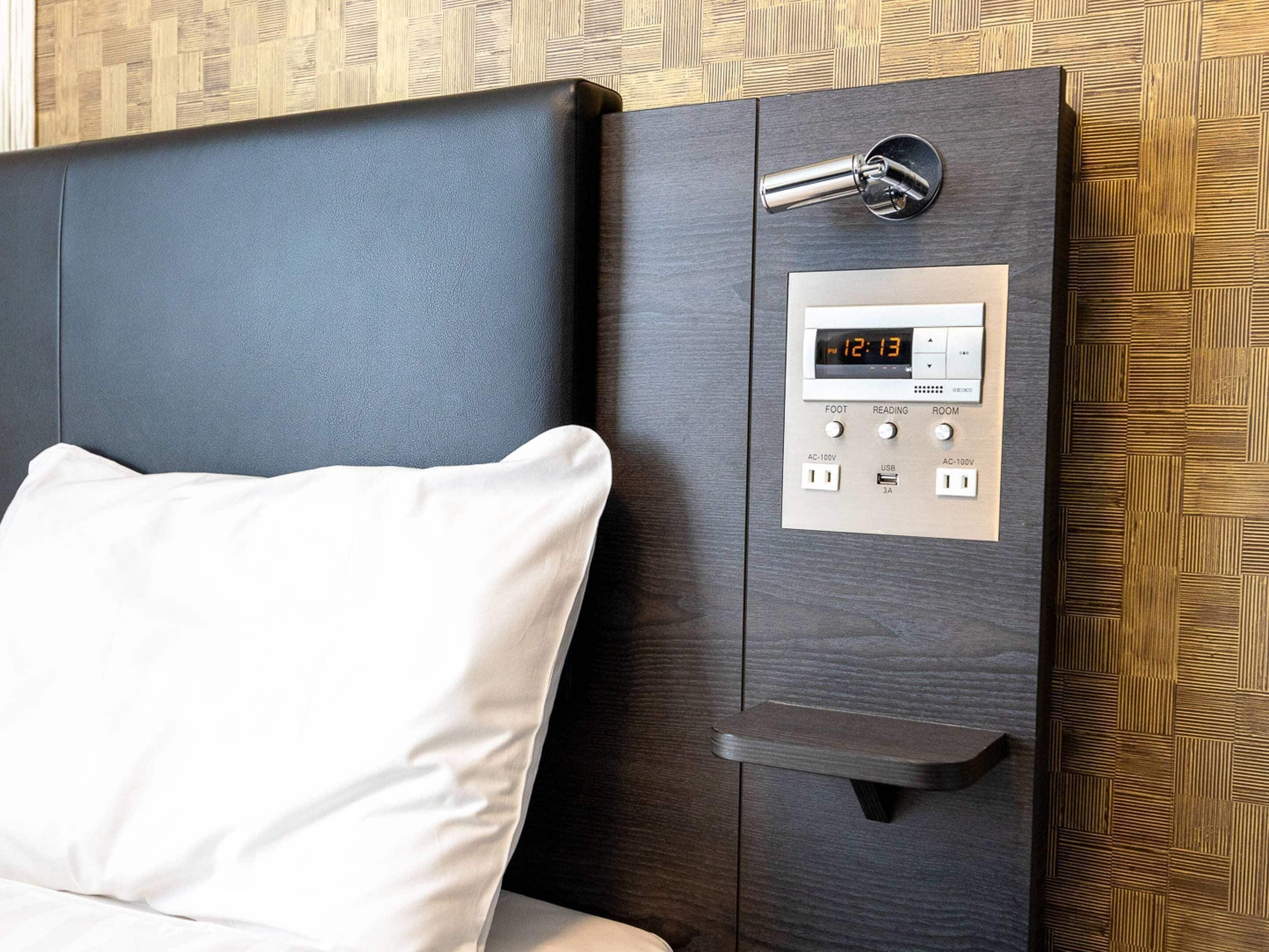 Una habitación moderna con cama, un reloj digital y un panel de control con botones para los pies, lectura y la habitación.