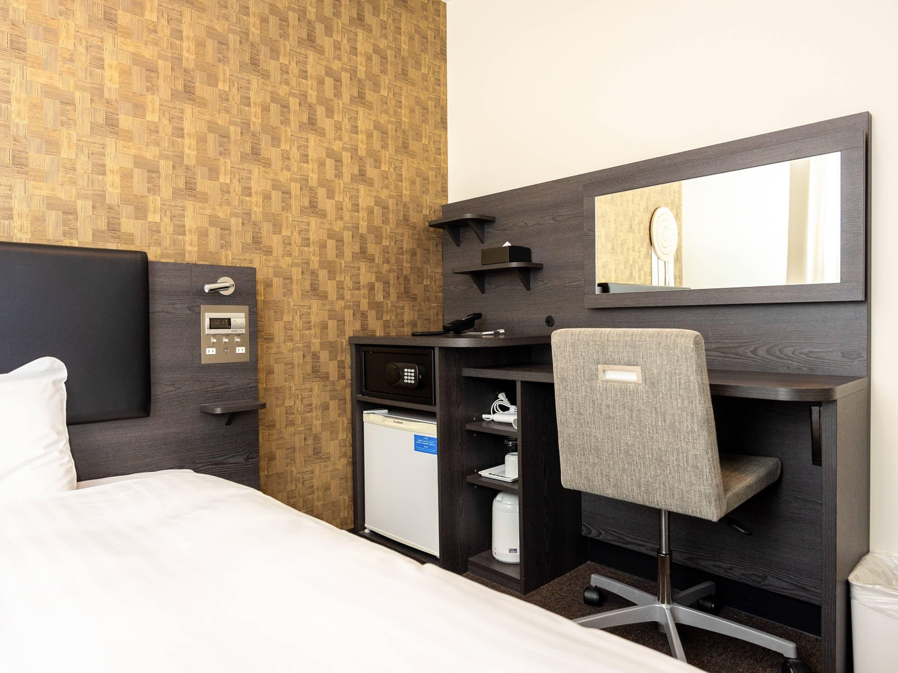 Ein Hotelzimmer mit einem Bett, einem Schreibtisch, einem Stuhl, einem kleinen Kühlschrank, einer Mikrowelle, einer Kaffeemaschine, einem Telefon, einer Bedieneinheit und einem Spiegel.