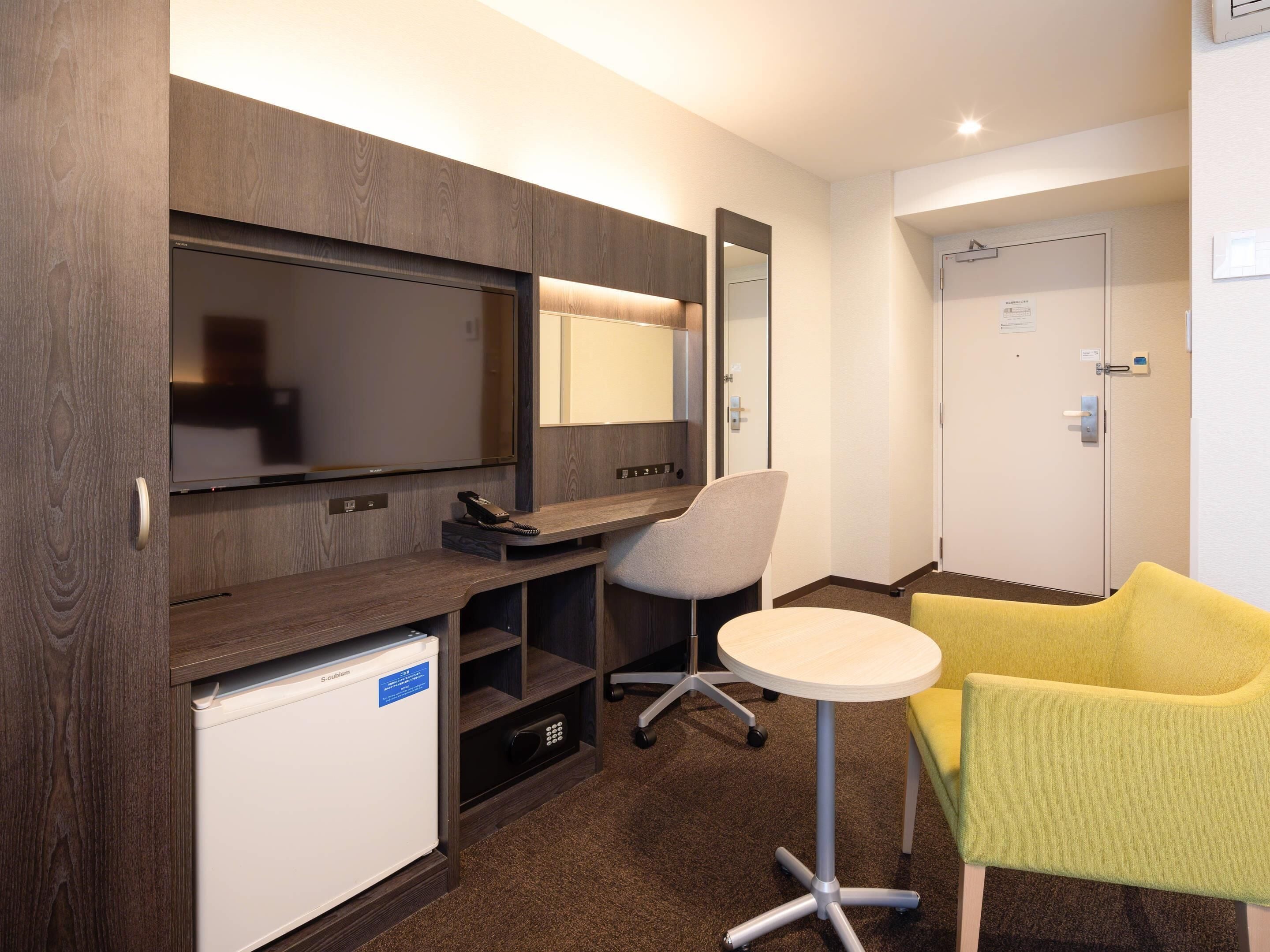 Ein Hotelzimmer mit Fernseher, kleinem Kühlschrank, Schreibtisch mit Stuhl und gelbem Sessel.