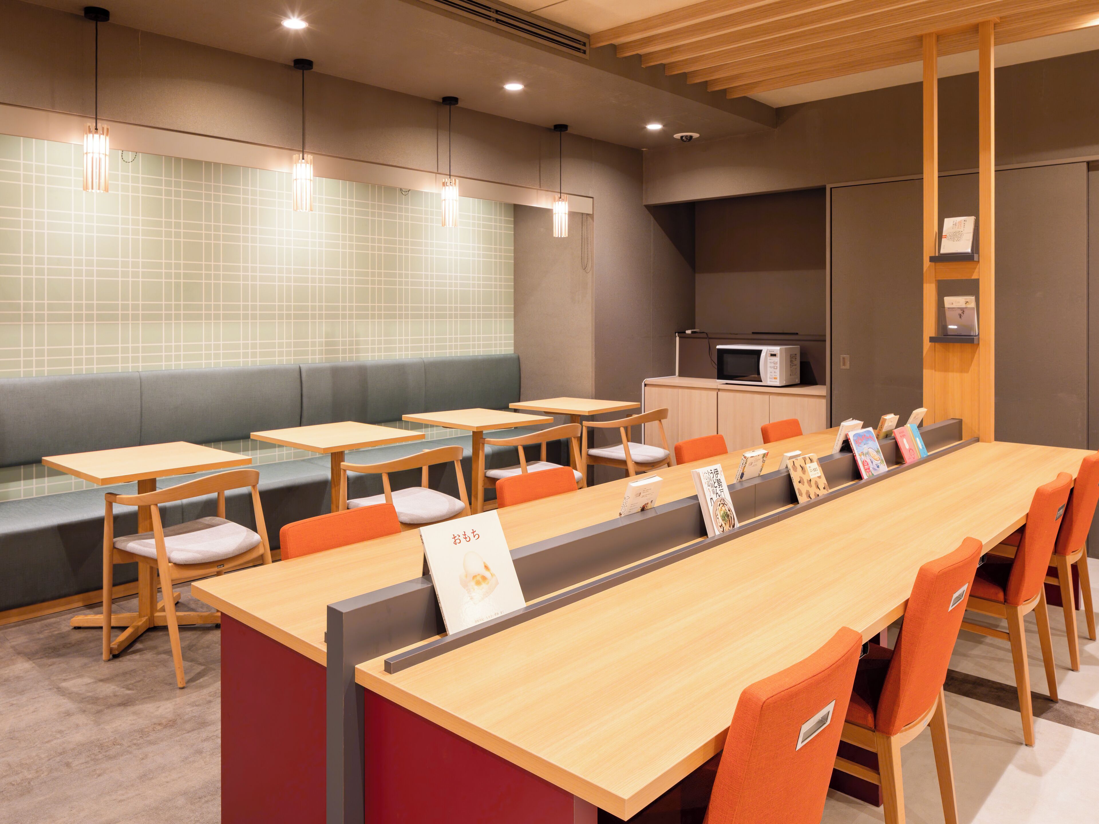 A modern cafe with wooden tables and orange chairs, a long bench, and a microwave on a shelf.