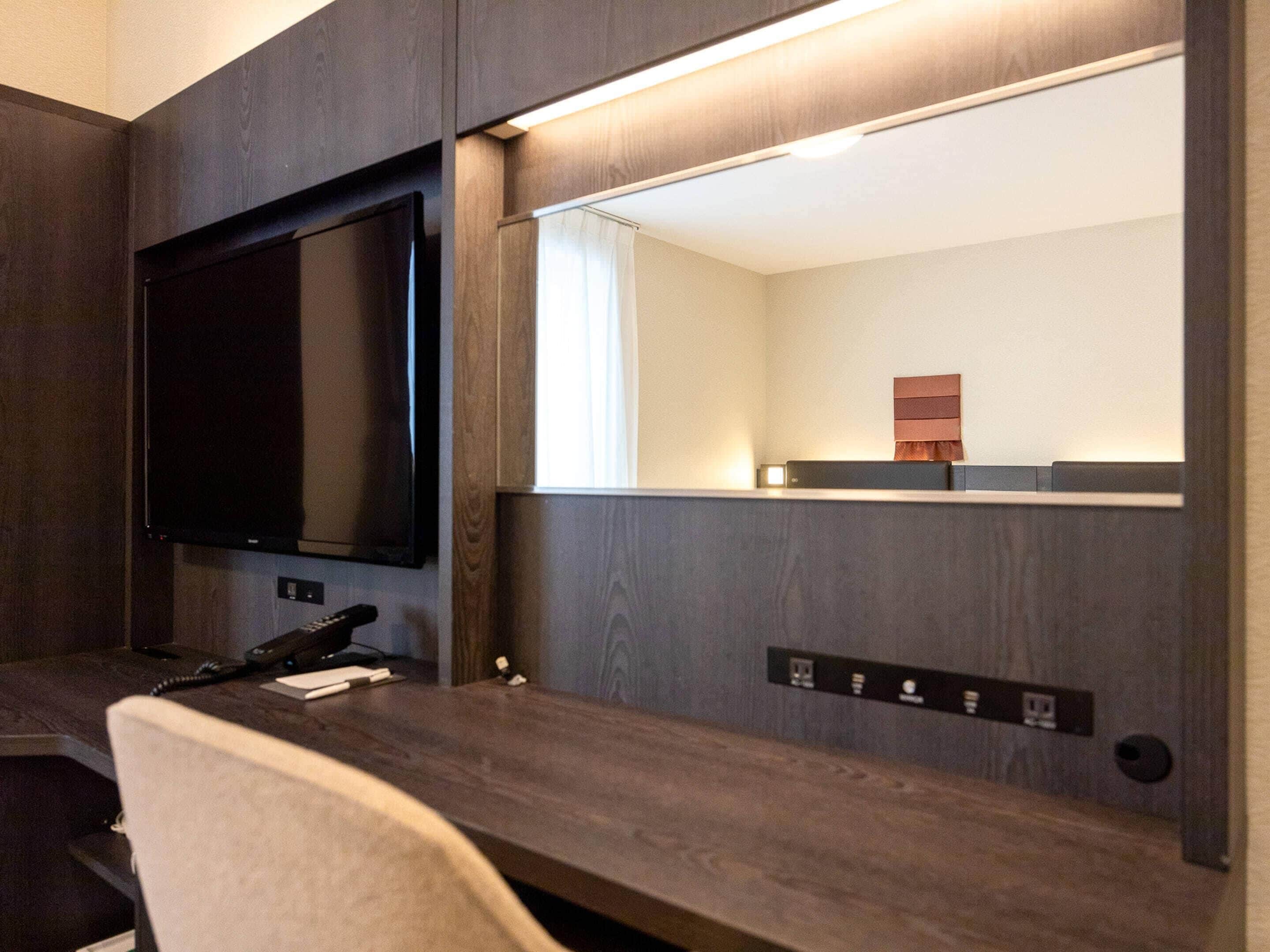 A hotel room with a flat-screen TV mounted on the wall, a desk with a telephone, and a power outlet.