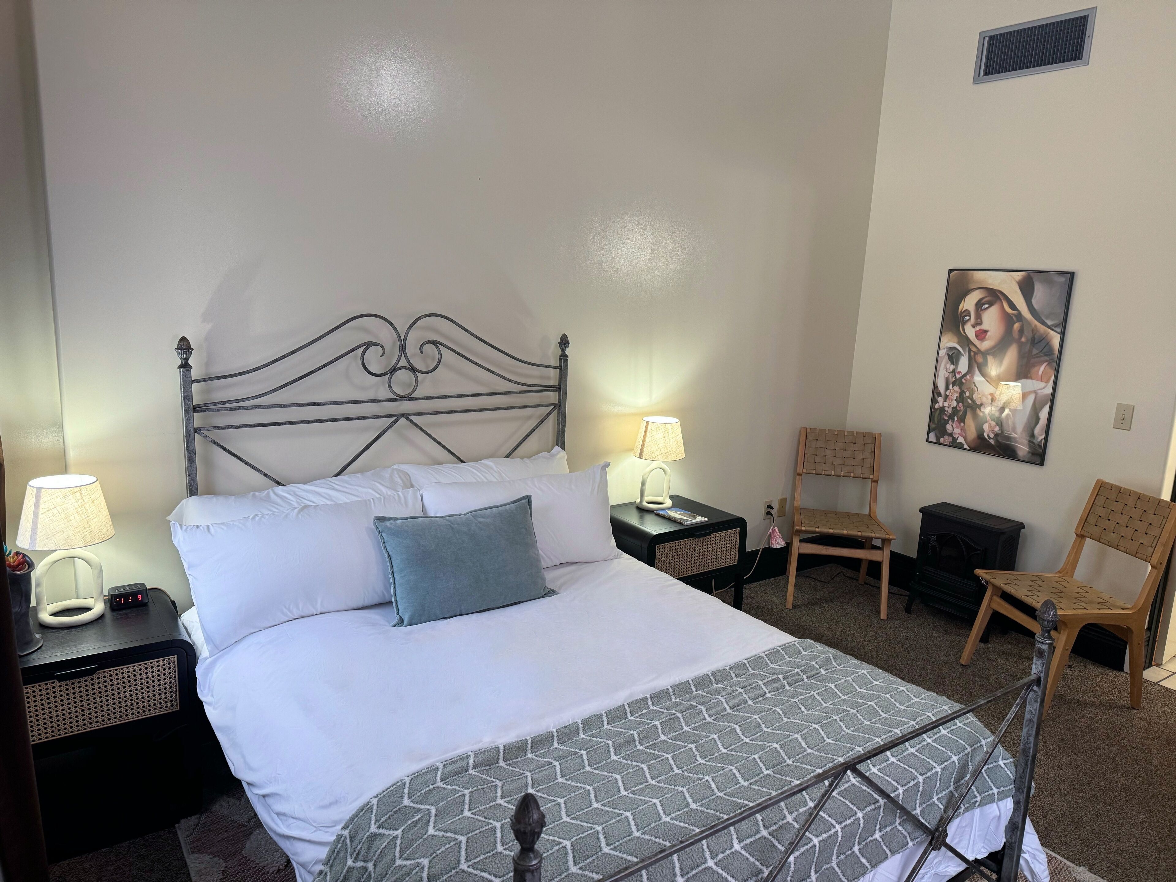 A neatly made bed with a metal headboard, a bedside table with a lamp, a chair, and a painting on the wall.