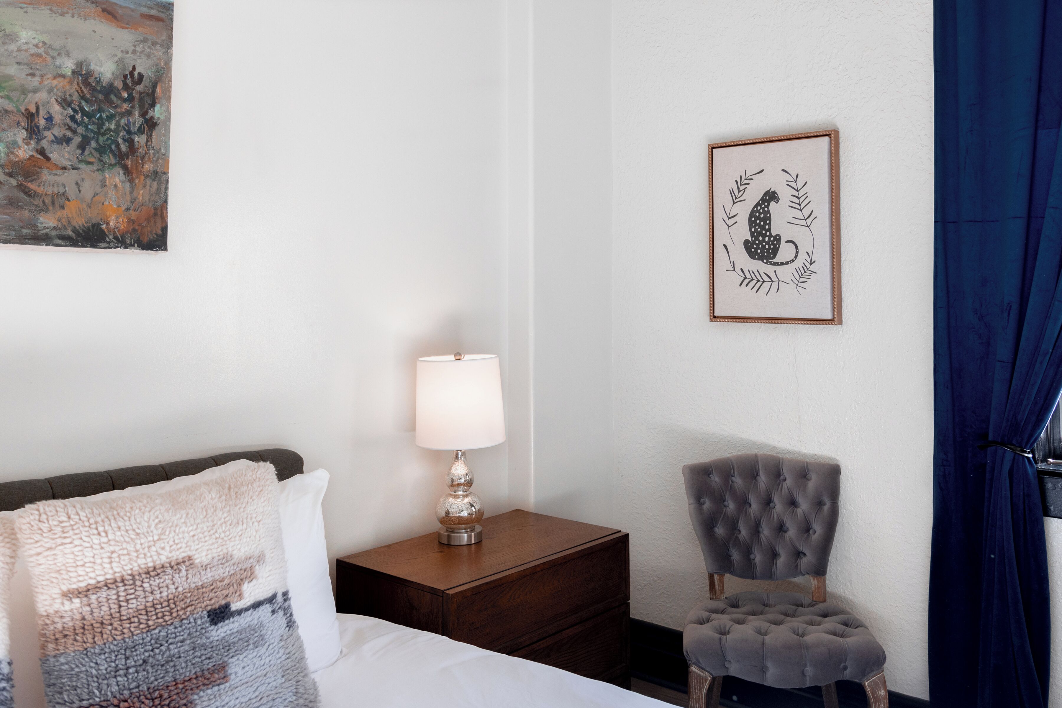 Habitación de hotel con cama, mesita de noche con lámpara, una obra de arte enmarco, una silla y cortina azul.