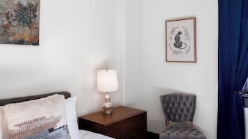 Habitación de hotel con cama, mesita de noche con lámpara, una obra de arte enmarco, una silla y cortina azul.