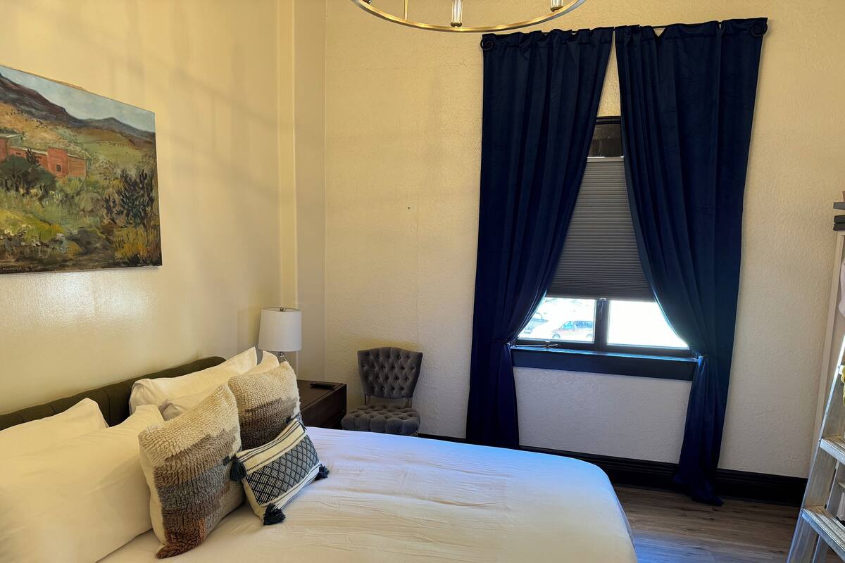 Une chambre d’hôtel avec un grand lit, un tableau au mur, une chaise et une fenêtre avec des rideaux foncés.