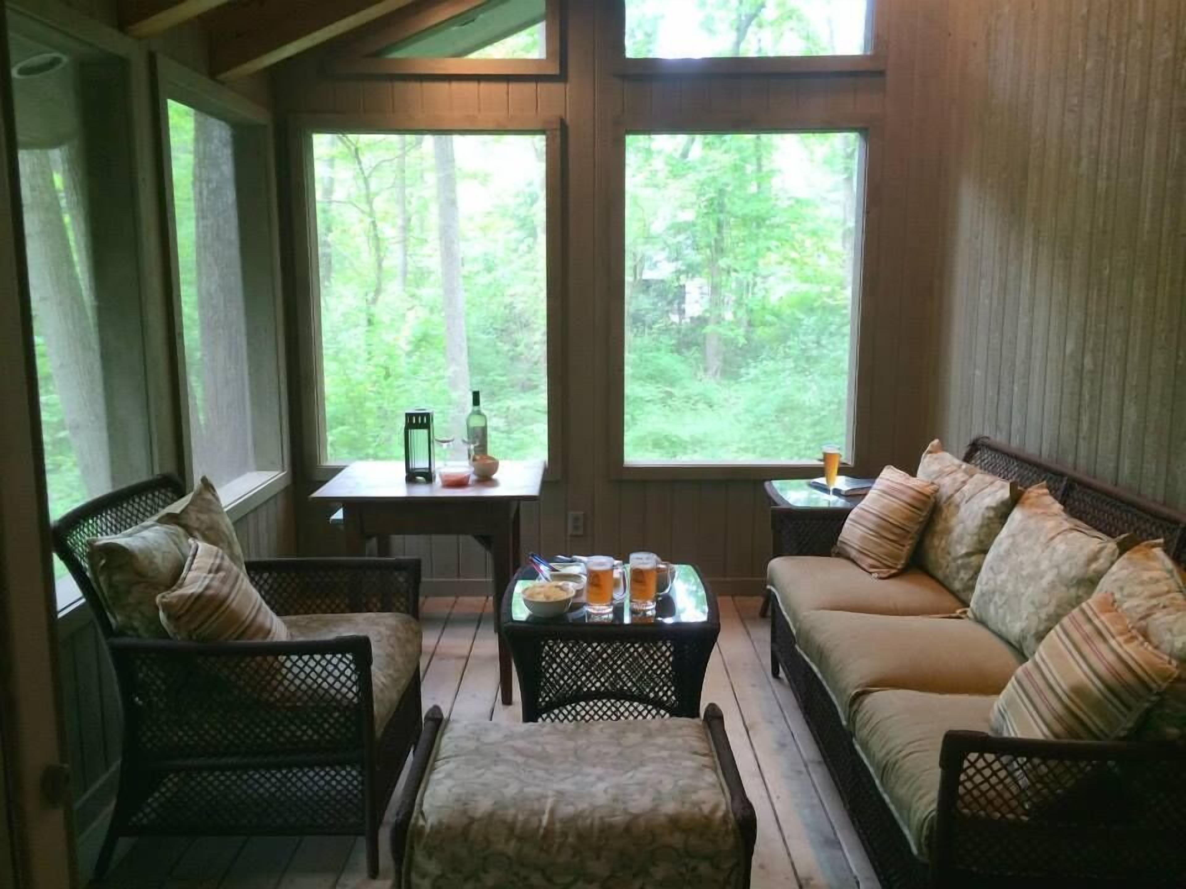 A cozy room with a sofa, chairs, and a table with drinks and a bottle. Large windows offer a view of the forest.