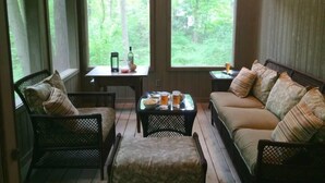Ein gemütlicher Raum mit einer Couch, Stühlen und einem Tisch mit Getränken und einer Flasche. Große Fenster bieten Blick auf den Wald.