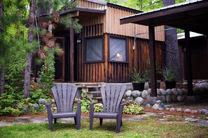 A wooden cabin with two Adirondack chairs in front, surrounded by a garden with rocks and plants. - Beautiful Waterfront Log Cottage with Dock, Beach & Sleep Cabin Near Mattawa (Mattawan)