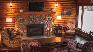 Interior acogedor de una cabaña de madera con chimenea de piedra, muebles de cuero y un amplio ventanal con vistas al exterior.