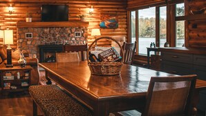 Interior de una cabaña de madera con chimenea de piedra, una mesa de comedor, sillas y un televisor montado en la pared.
