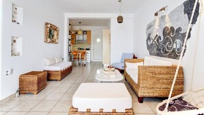 Ein modernes Wohnzimmer mit Rattanmöbeln, einem weißen Sofa, einem Essbereich mit Tisch und Stühlen sowie einer Küche im Hintergrund.